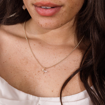 A close-up of a woman wearing a delicate gold bead chain necklace with a small dragonfly pendant. The necklace is shown on her neck, with her dark hair and white top visible.