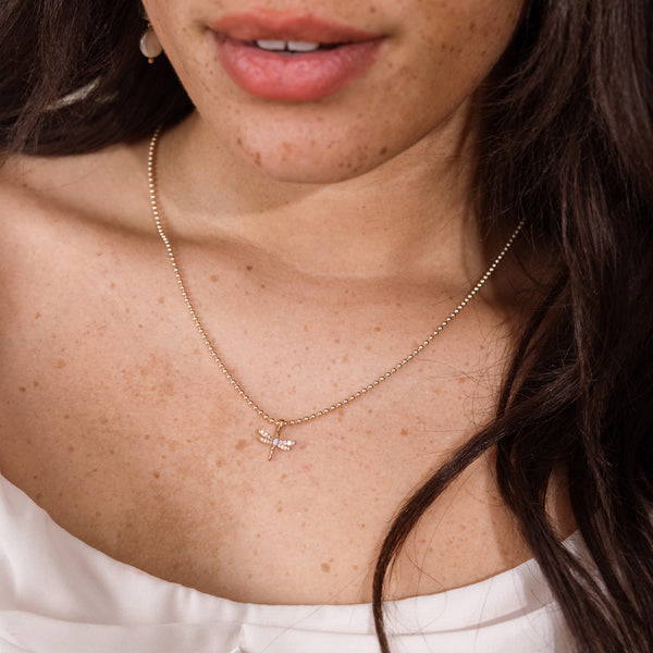 A close-up of a woman wearing a delicate gold bead chain necklace with a small dragonfly pendant. The necklace is shown on her neck, with her dark hair and white top visible.