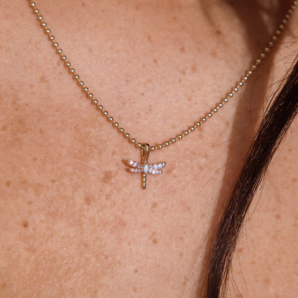 A close-up of a delicate gold ball-chain necklace with a small dragonfly pendant, worn on a person's neck. The dragonfly pendant features clear rhinestones on its wings and an opal stone at the center.