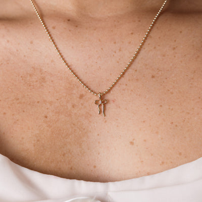 A delicate gold beaded chain necklace with a small bow-shaped pendant, shown worn on a person's neck and upper chest.