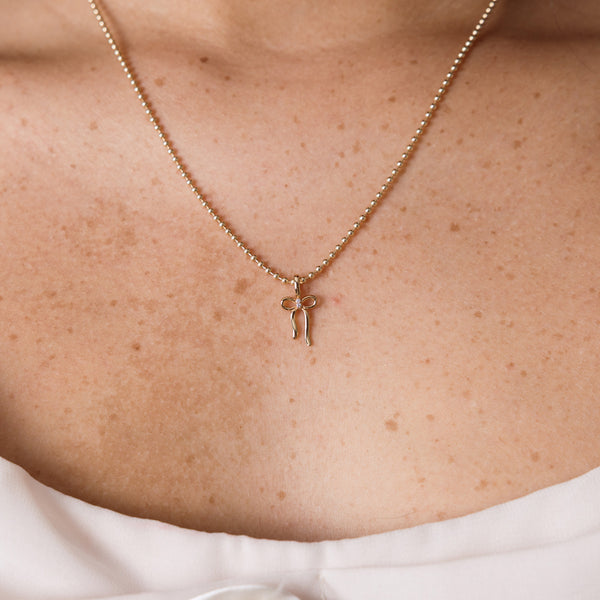 A delicate gold beaded chain necklace with a small bow-shaped pendant, shown worn on a person's neck and upper chest.