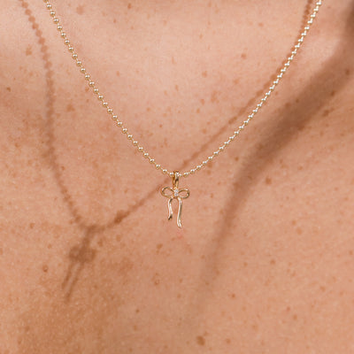 A delicate gold bead chain necklace with a small bow-shaped pendant, shown worn on a person's neck against a warm skin tone background.