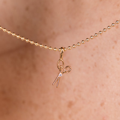 A close-up of a delicate gold bead chain necklace worn against skin, featuring a small scissors pendant with a tiny clear gemstone at the center.