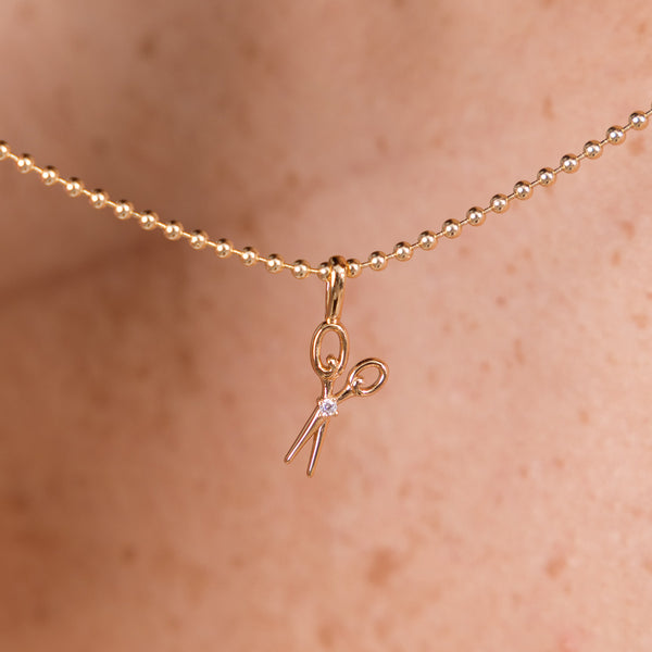 A close-up of a delicate gold bead chain necklace worn against skin, featuring a small scissors pendant with a tiny clear gemstone at the center.