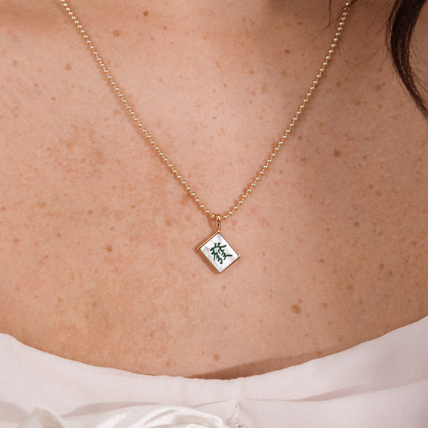 A close-up of a person's chest wearing a delicate gold bead chain necklace with a small square mahjong tile pendant. The pendant has a white center with a green Chinese character and a gold border.