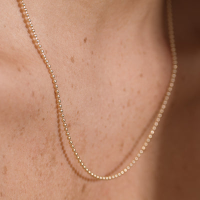A close-up of a delicate gold beaded chain necklace worn on a person's neck, showing the fine metallic links against warm skin tones.