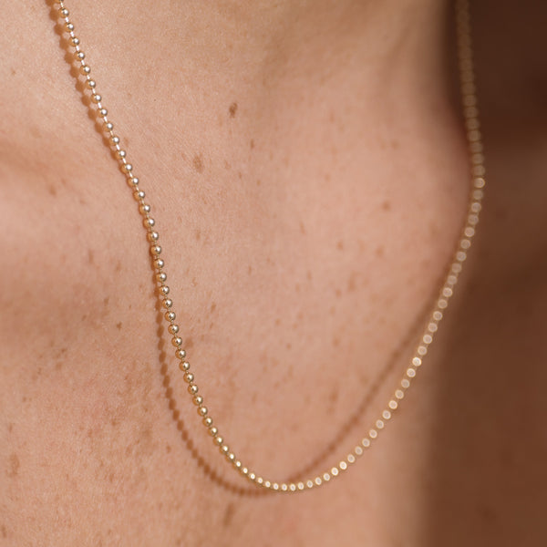 A close-up of a delicate gold beaded chain necklace worn on a person's neck, showing the fine metallic links against warm skin tones.