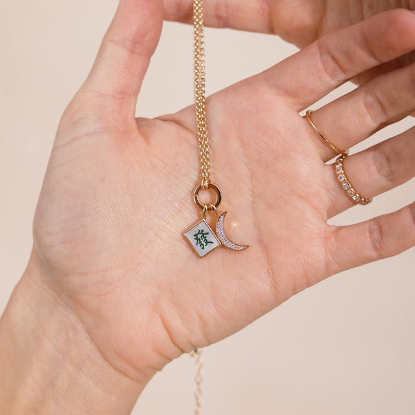 A gold chain necklace with a round link lock holding a square mahjong tile and crescent moon pendant displayed on a person's palm. The square pendant features a green and white Japanese-style pattern, and the chain and moon are gold-toned.