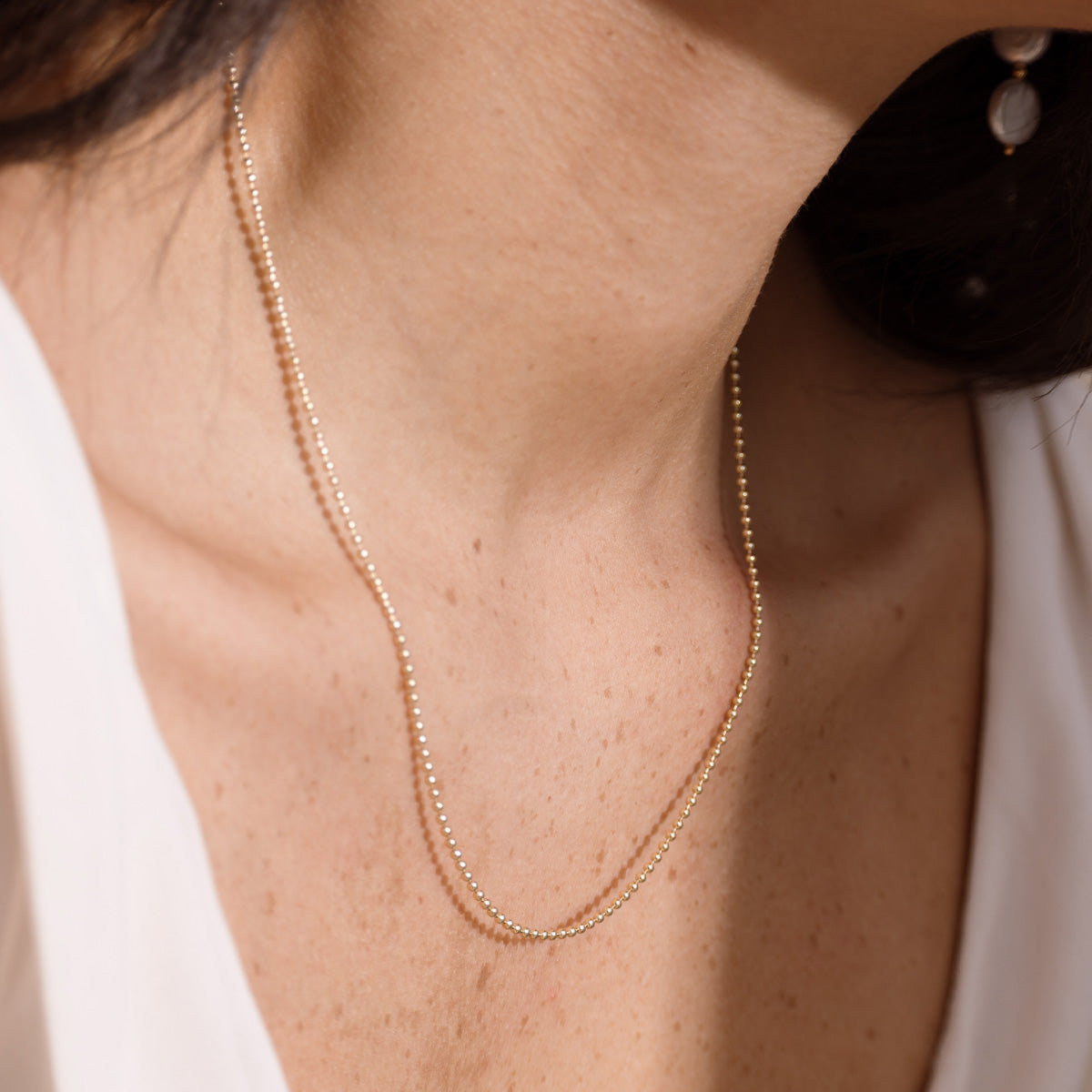 A delicate gold bead chain necklace worn on a model's neck, shown in a close-up lifestyle shot with a white top.
