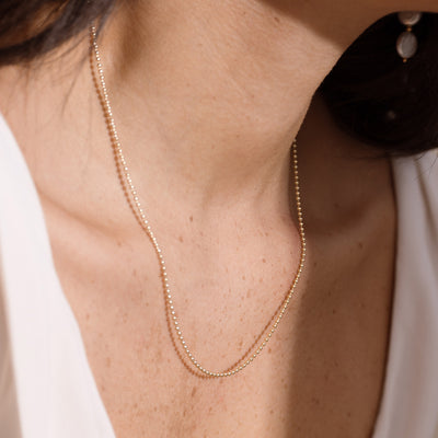 A delicate gold bead chain necklace worn on a model's neck, shown in a close-up lifestyle shot with a white top.