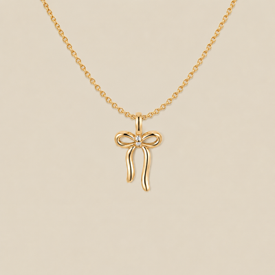A gold chain necklace featuring a bow-shaped pendant with a small clear gemstone at the center, displayed against a solid beige background.