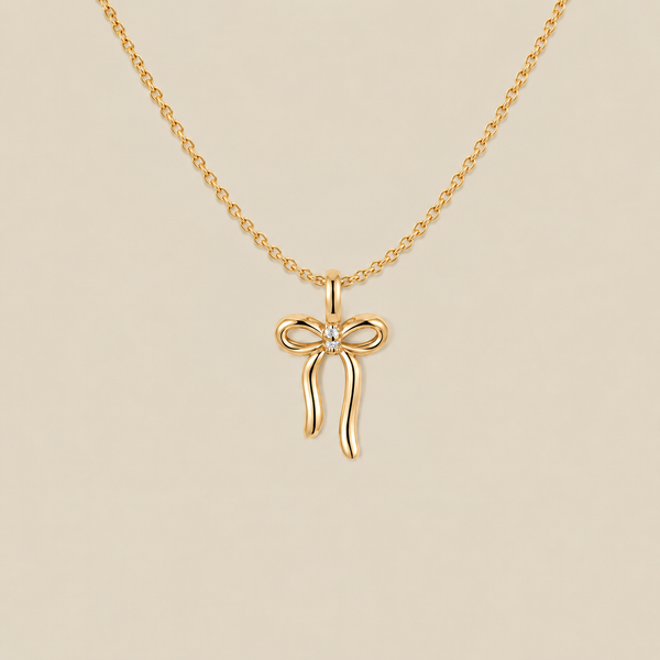 A gold chain necklace featuring a bow-shaped pendant with a small clear gemstone at the center, displayed against a solid beige background.