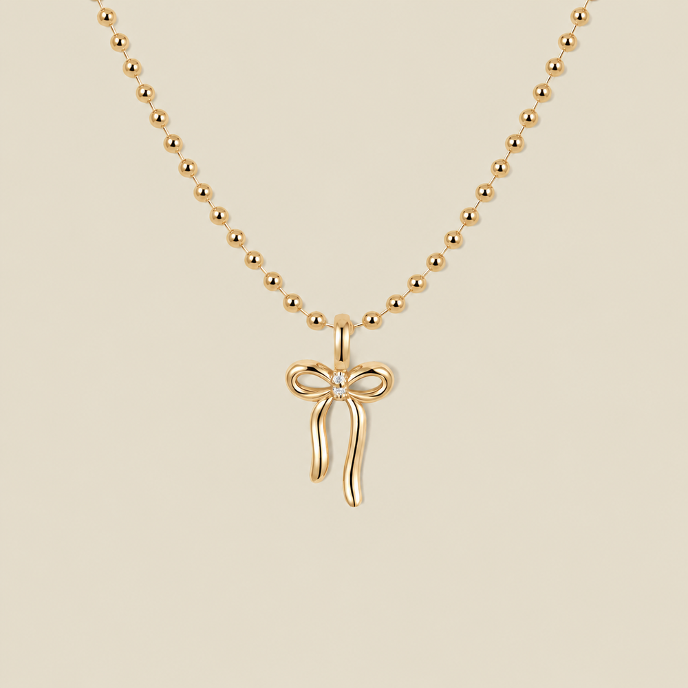 A gold ball chain necklace featuring a small bow pendant with a clear gemstone in the center, displayed against a solid beige background.