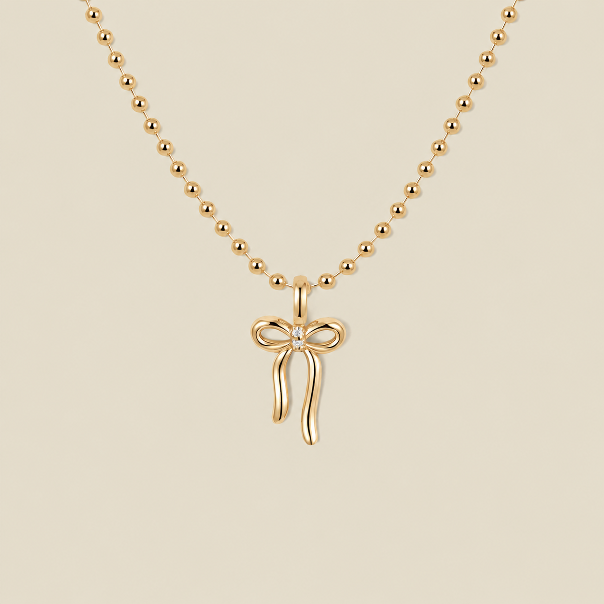 A gold ball chain necklace featuring a small bow pendant with a clear gemstone in the center, displayed against a solid beige background.