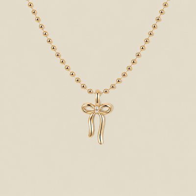 A gold ball chain necklace featuring a small bow pendant with a clear gemstone in the center, displayed against a solid beige background.