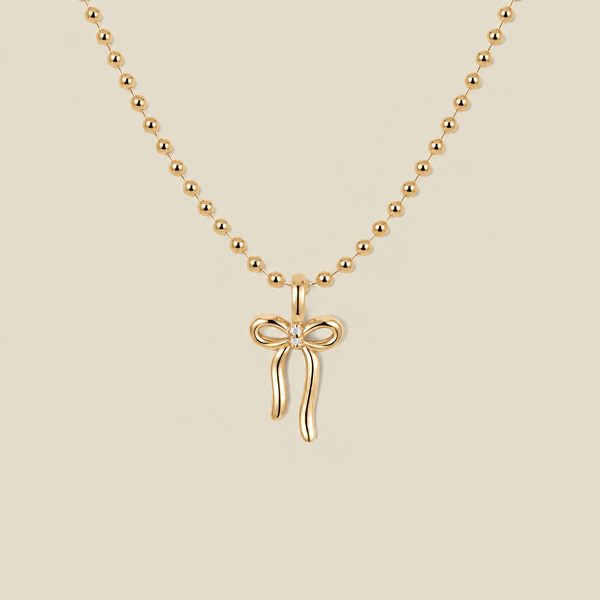A gold ball chain necklace featuring a small bow pendant with a clear gemstone in the center, displayed against a solid beige background.