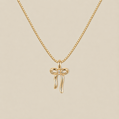 A delicate gold box chain necklace featuring a small bow-shaped pendant with a tiny clear gemstone at the center, displayed against a solid beige background.
