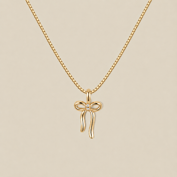 A delicate gold box chain necklace featuring a small bow-shaped pendant with a tiny clear gemstone at the center, displayed against a solid beige background.