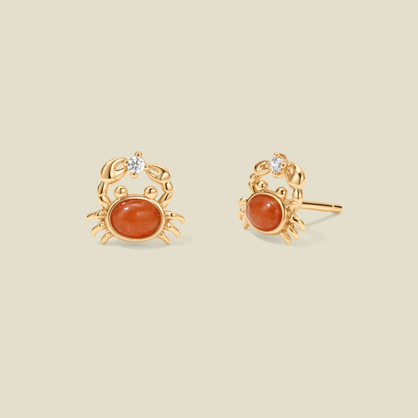 A pair of gold crab stud earrings with oval orange-red  stones and a small clear stone on top, shown on a plain warm beige background.