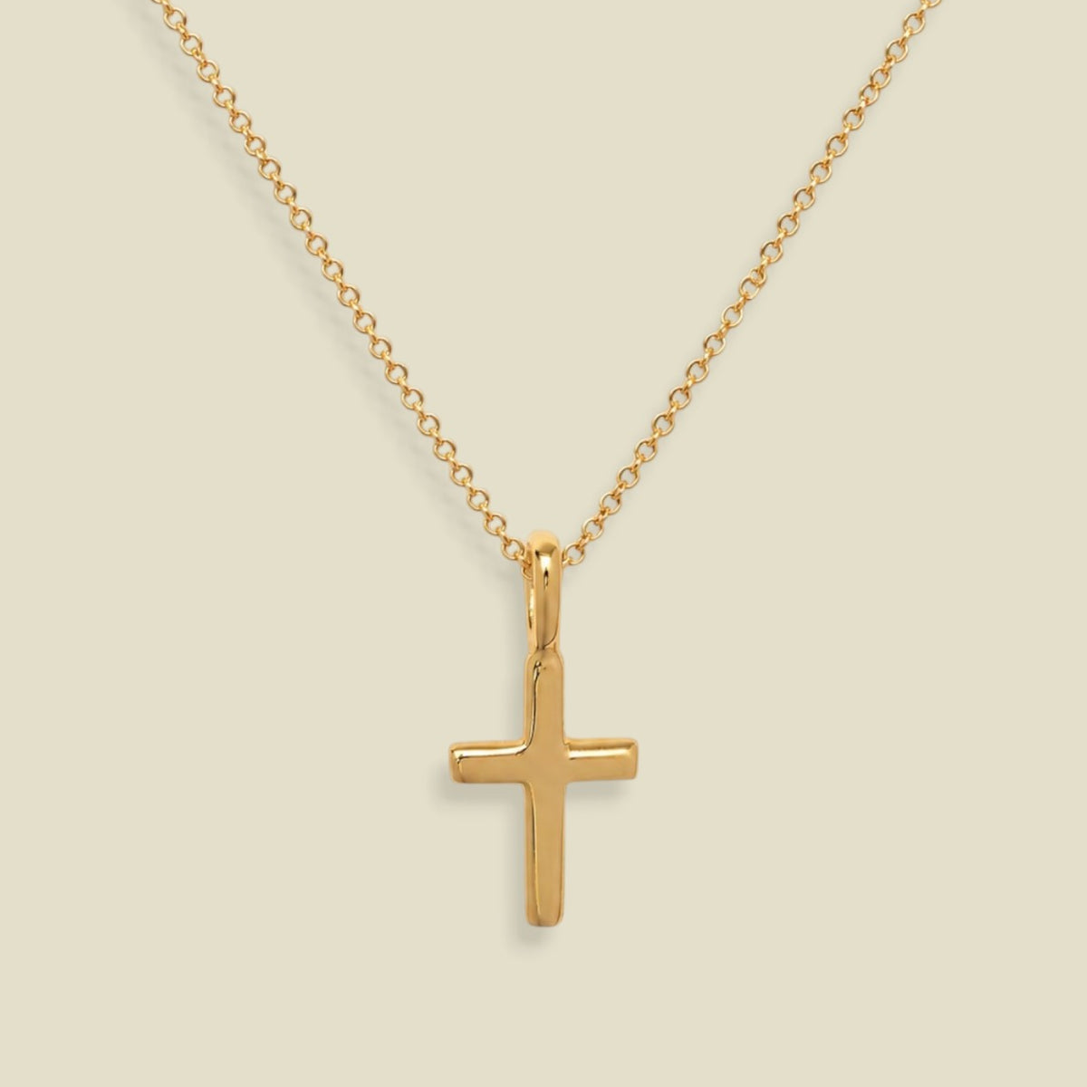 A dainty gold chain with a small gold cross charm, set against a solid soft beige background.
