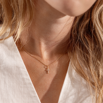 Close-up of a person wearing a delicate necklace with a cross pendant with clear stones, set against a neutral background.