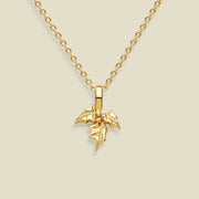 A gold holly flower pendant necklace on a delicate gold chain, presented against a solid light beige background.
