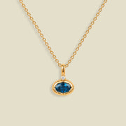 A gold chain necklace with a pendant featuring an oval cut blue gemstone in a gold bezel setting, accented by a small clear stone on the top, set against a solid light beige background.