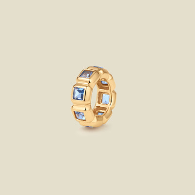 A gold ring charm with multiple bezel set square-cut light blue gemstones set side by side to create the band, shown on a solid beige background.