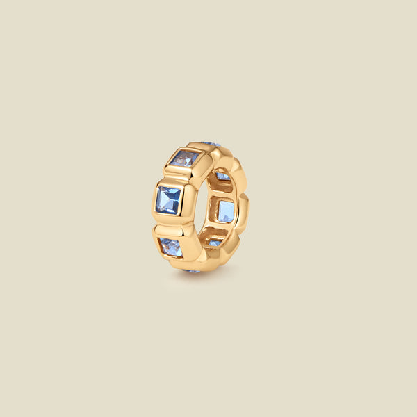 A gold ring charm with multiple bezel set square-cut light blue gemstones set side by side to create the band, shown on a solid beige background.