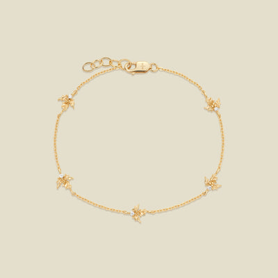 A delicate gold chain bracelet with five small gold holly charms, each accented with a tiny clear crystal, displayed on a solid light beige background.