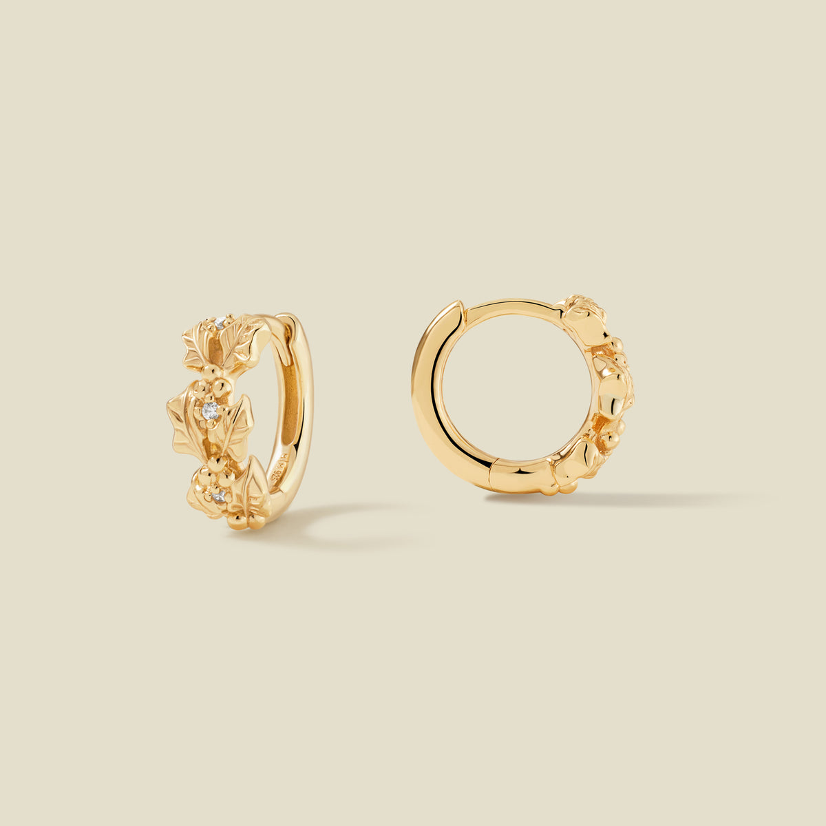 A pair of gold hoop earrings adorned with textured holly flowers stacked along the hoop, with scattered small clear accent stones, set against a solid beige background.