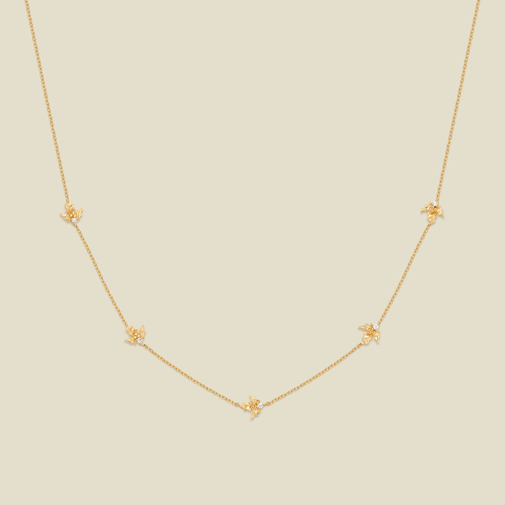 A delicate gold chain necklace featuring five small, evenly spaced textured holly flower motifs set with tiny clear stones, displayed on a solid light beige background.