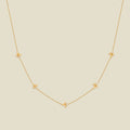 A delicate gold chain necklace featuring five small, evenly spaced textured holly flower motifs set with tiny clear stones, displayed on a solid light beige background.