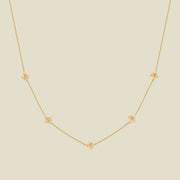 A delicate gold chain necklace featuring five small, evenly spaced textured holly flower motifs set with tiny clear stones, displayed on a solid light beige background.