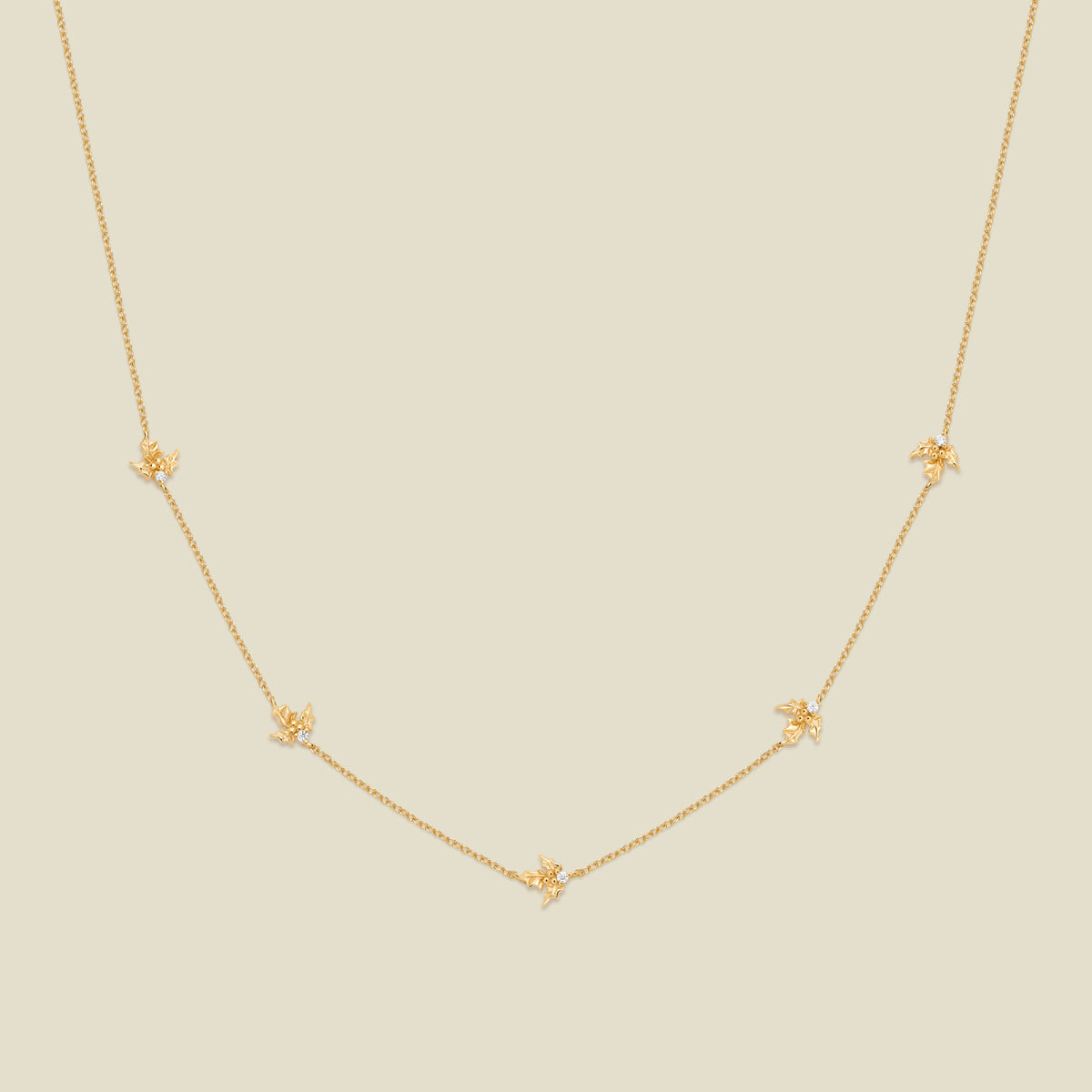 A delicate gold chain necklace featuring five small, evenly spaced textured holly flower motifs set with tiny clear stones, displayed on a solid light beige background.