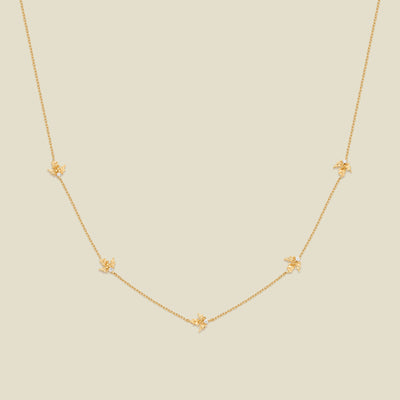 A delicate gold chain necklace featuring five small, evenly spaced textured holly flower motifs set with tiny clear stones, displayed on a solid light beige background.