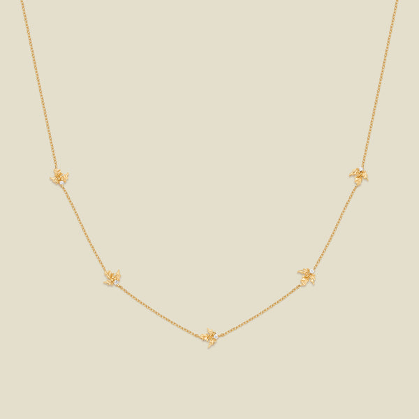 A delicate gold chain necklace featuring five small, evenly spaced textured holly flower motifs set with tiny clear stones, displayed on a solid light beige background.