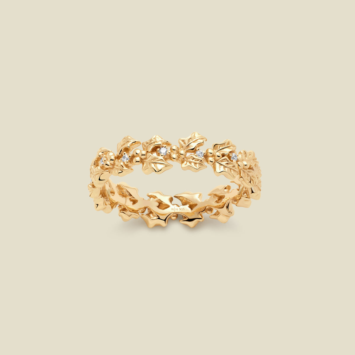 A gold floral-etched ring band with holly flowers and small clear gemstones, shown on a plain warm beige background. 