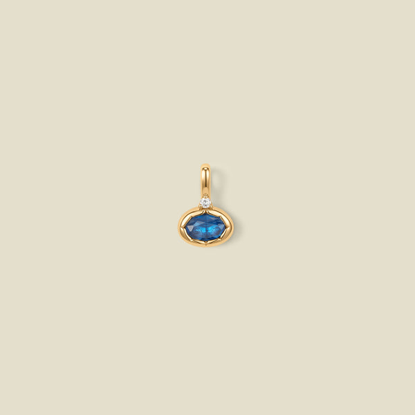 A gold oval pendant charm with a blue bezel-set oval gemstone and a clear accent stone on top, set against a solid beige background.