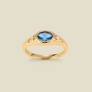 A gold signet ring with a blue center oval bezel-set gemstone, and two small clear accent stones on either side, set against a solid beige background.