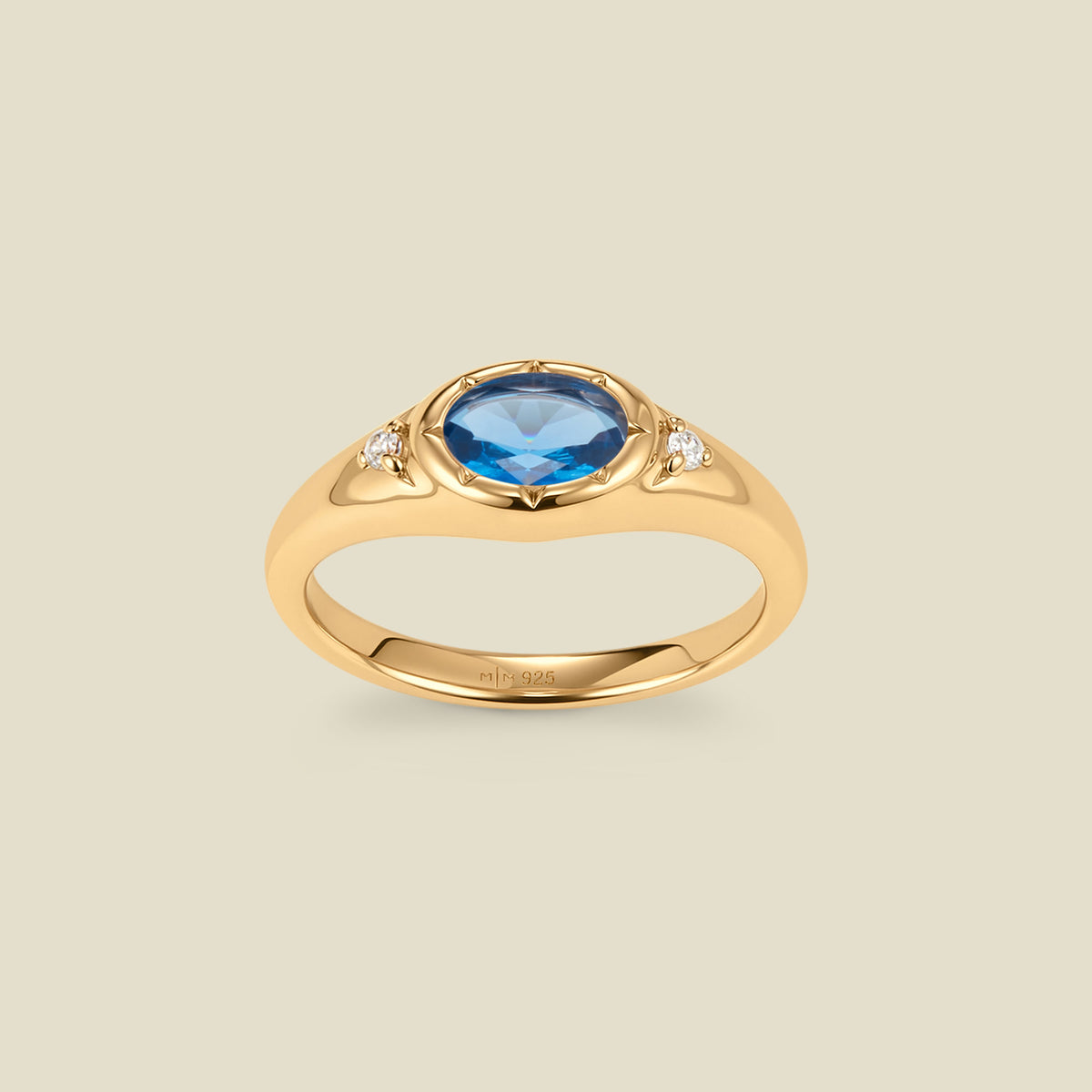 A gold signet ring with a blue center oval bezel-set gemstone, and two small clear accent stones on either side, set against a solid beige background.