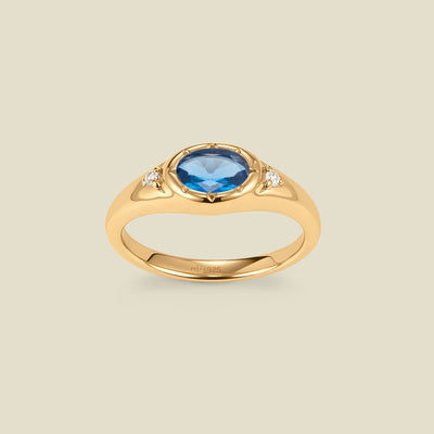 A gold signet ring with a blue center oval bezel-set gemstone, and two small clear accent stones on either side, set against a solid beige background.
