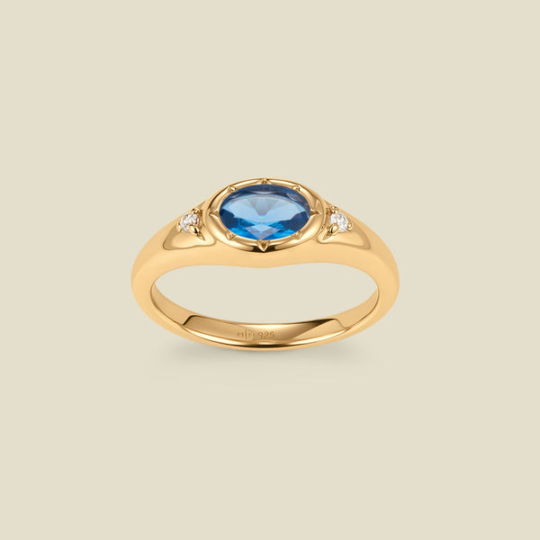 A gold signet ring with a blue center oval bezel-set gemstone, and two small clear accent stones on either side, set against a solid beige background.