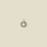 A circular gold pendant featuring blue baguette-cut stones around a hollow center, displayed against a solid light beige background.