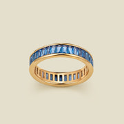 A gold eternity ring featuring a continuous channel setting of baguette-cut blue gemstones, shown on a solid light beige background.
