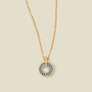 A gold chain necklace with a circular pendant featuring blue baguette-cut stones around a hollow center, displayed against a solid light beige background.