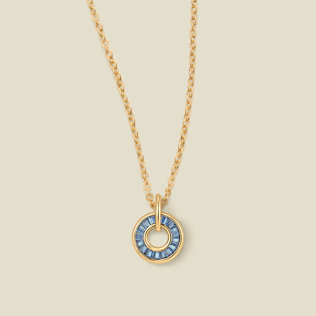 A gold chain necklace with a circular pendant featuring blue baguette-cut stones around a hollow center, displayed against a solid light beige background.
