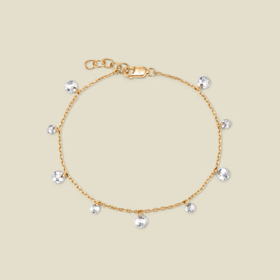 A dainty gold chain bracelet with multiple clear round crystals dangling along it, shown against a solid beige background.