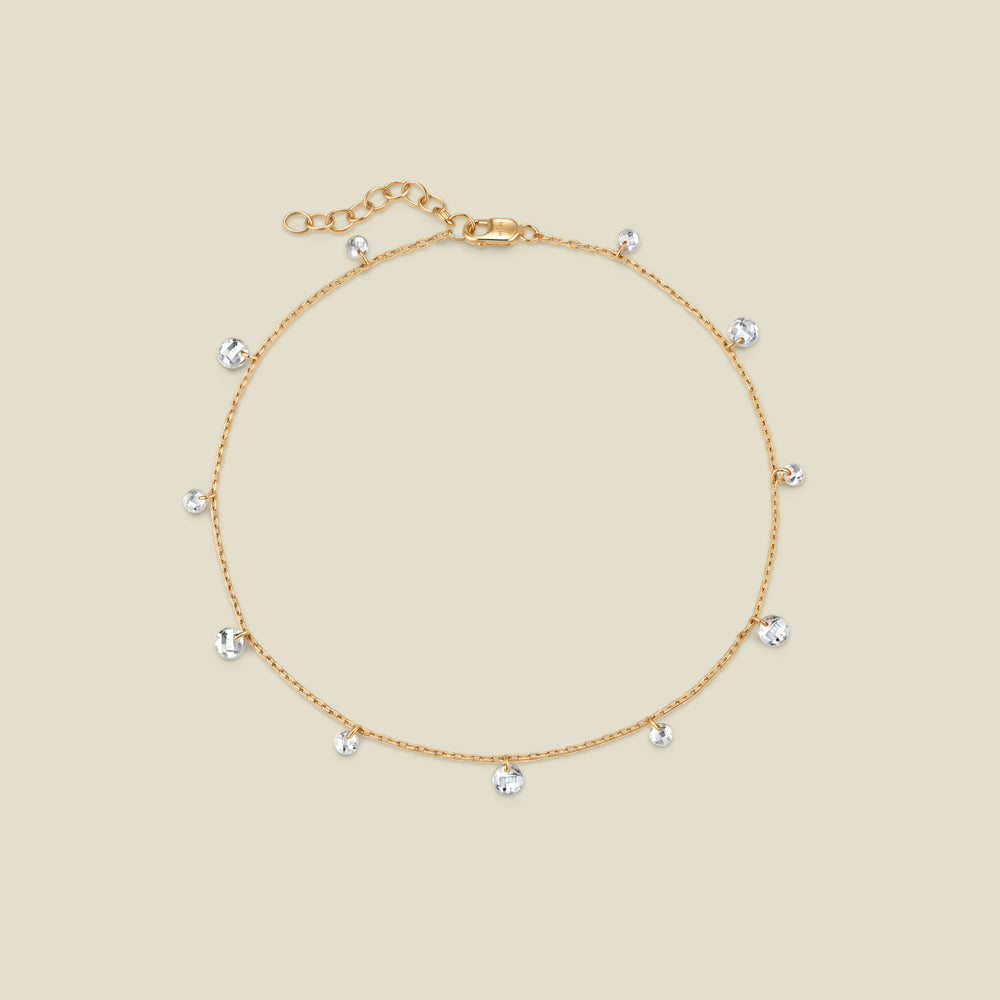 A dainty gold chain anklet with clear round crystals dangling along it, on a solid beige background.