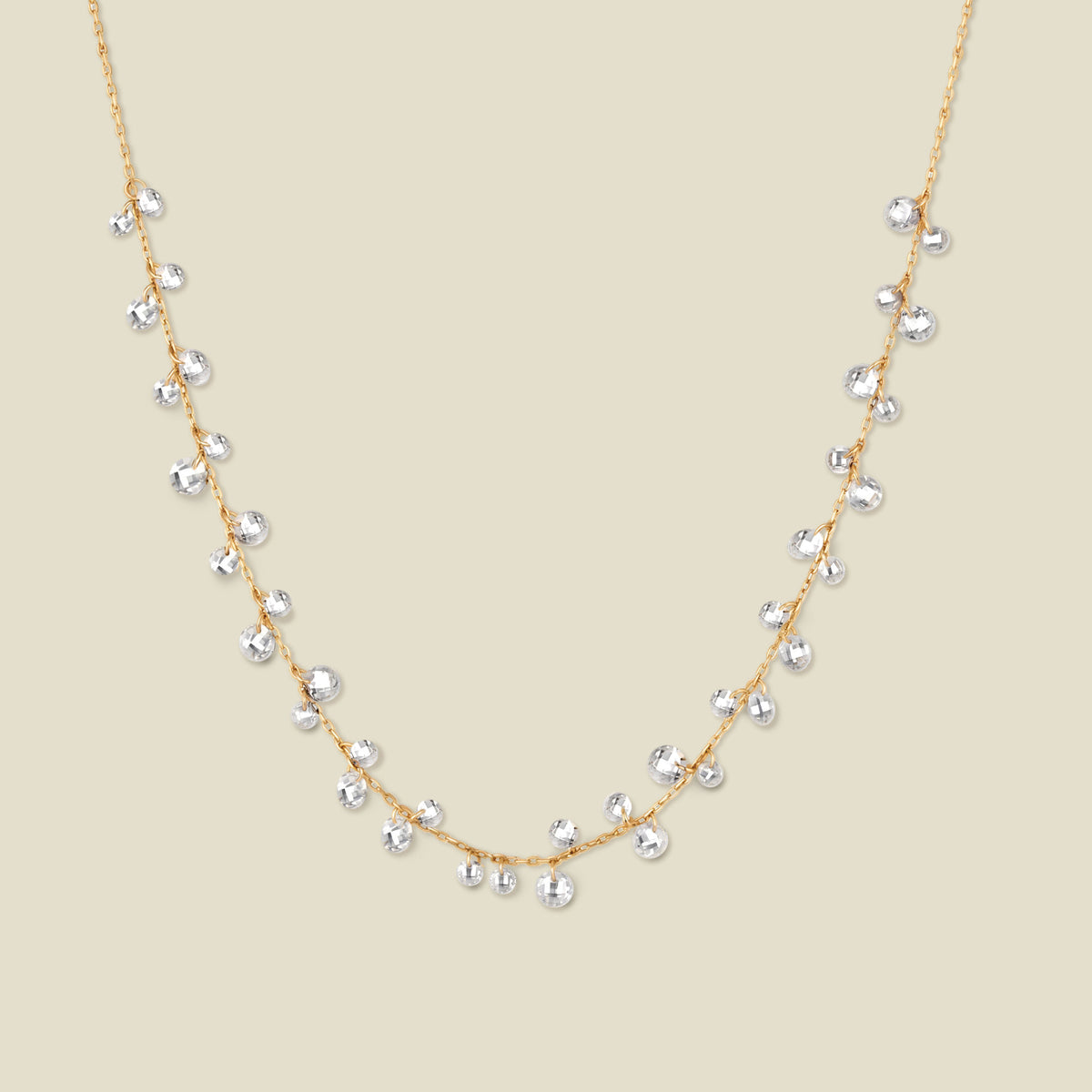 A dainty gold chain necklace with clusters of clear, round crystals dangling off, shown against a solid beige background.